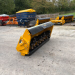 Yellow agricultural equipment with black toothed rollers, mounted on a frame in a yard with other farm machinery and pallets nearby.