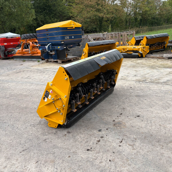 Yellow agricultural equipment with black toothed rollers, mounted on a frame in a yard with other farm machinery and pallets nearby.