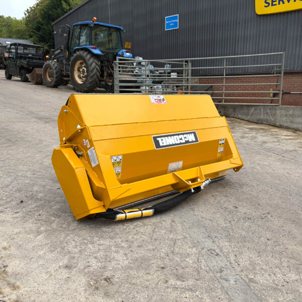 Yellow agricultural attachment in the foreground, with blue tractors and a service building in the background at an outdoor equipment yard.