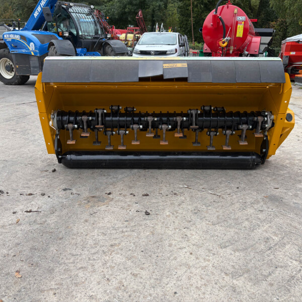Front view of a yellow agricultural machine with black rollers, parked in a yard with tractors and equipment in the background.