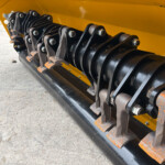 Close-up of a belt and roller mechanism on a yellow industrial machine, showing multiple black metal strips and bolts.
