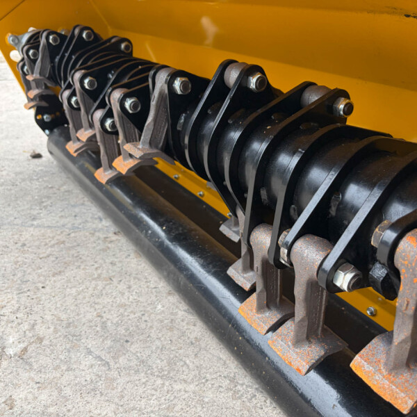 Close-up of a belt and roller mechanism on a yellow industrial machine, showing multiple black metal strips and bolts.