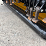 Close-up of industrial machinery track with rusted pins and a yellow frame on a black base, resting on concrete ground.