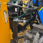 Close-up of orange and blue heavy machinery frame with hydraulic hoses, metal linkages, and muddy tires visible.