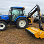 Blue New Holland tractor with front loader at an outdoor machinery show, yellow attachments in the foreground.