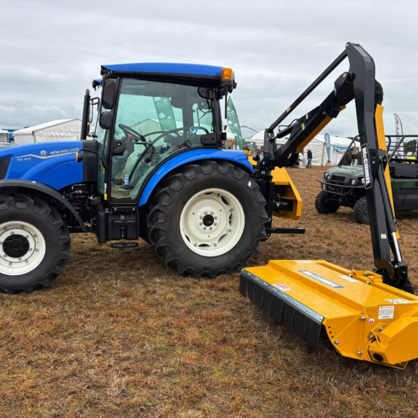 Blue New Holland tractor with front loader at an outdoor machinery show, yellow attachments in the foreground.