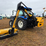 Yellow front-mounted farming attachment on a blue tractor at an outdoor machinery expo, with tents and people in the background.