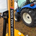 Blue tractor mounted on a yellow lifting device at an outdoor equipment show, with rain droplets on the surfaces and a muddy ground.