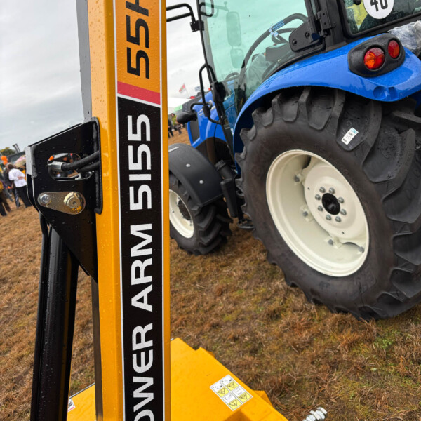 Blue tractor mounted on a yellow lifting device at an outdoor equipment show, with rain droplets on the surfaces and a muddy ground.