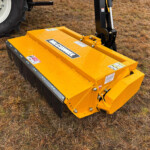 Yellow front-mounted tractor implement with hydraulic arm and warning labels, sitting on dry grass field (mower/attachment).