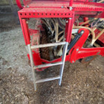 Red farm implement with perforated metal platform, ladder steps, and multiple hoses on a dirt floor.