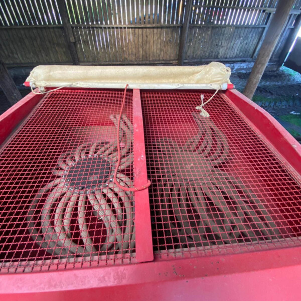 Red industrial machine with a red metal frame and a perforated mesh top, exposing two large cooling fans beneath, under a slatted wooden roof.