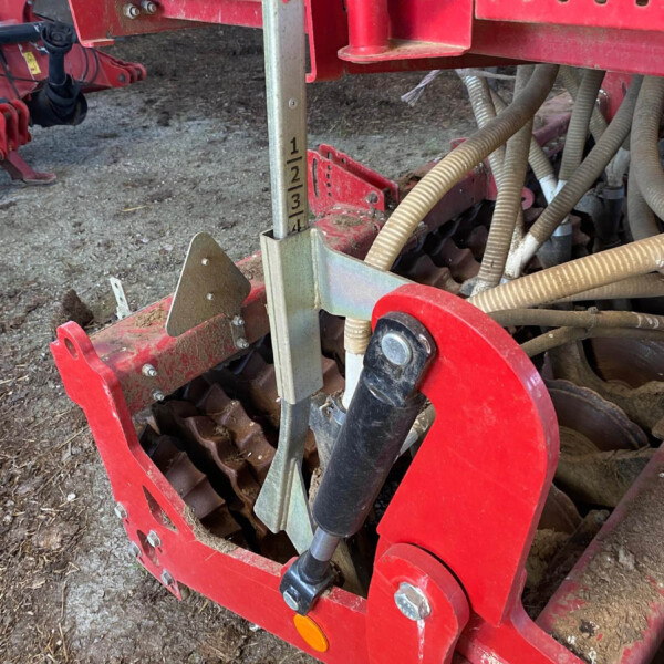 Close-up of a red farm implement showing a vertical metal depth gauge marked 1–4, with bolts, brackets and beige hoses nearby.
