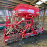 Red Moore uni-drill seed drill with multiple hoses, stored in a wooden shed.