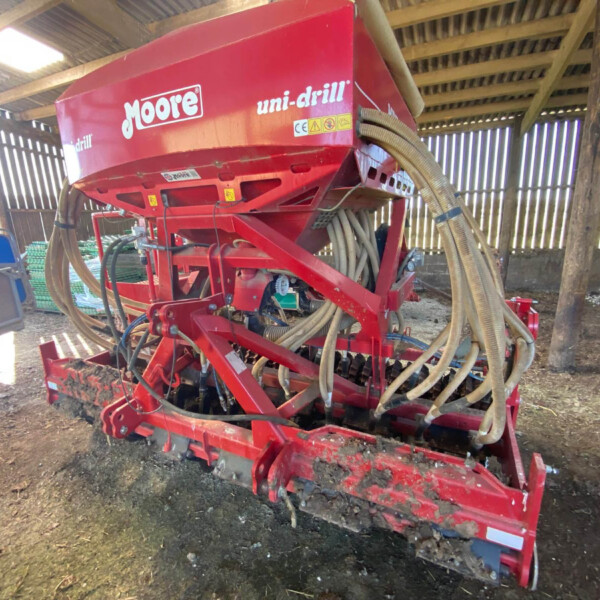 Red Moore uni-drill seed drill in a shed with hoses and metal frame, used for planting.