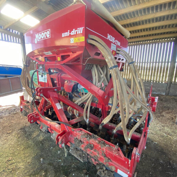 Red Moore uni-drill seed drill with numerous hoses attached, stored inside a wooden-shed barn.