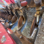 Red agricultural machine with multiple round disc blades and metal bolts, set beside a dirt ground.
