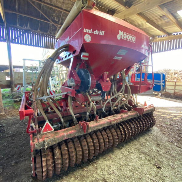 Red Moore Uni-Drill seed drill inside a shed with hoses and metal framework visible, ready for agricultural use.