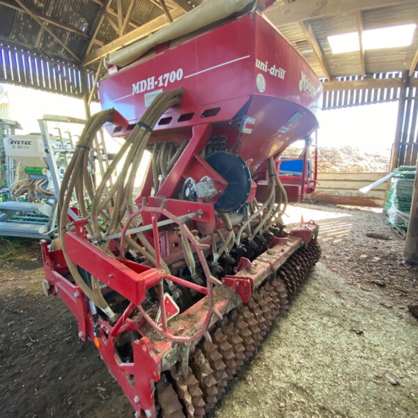 Red MDH-1700 uni-drill seed drill in a shed, showing multiple hoses and a row of spiked rollers for planting.