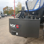 Front view of a blue tractor with a large black MX Multimass 1200 front weight and rugged tires in a yard.