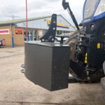 Close-up of a large industrial machine's counterweight and lifting arm in a service yard, with a building and signs in the background.