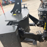 Close-up of a heavy machinery hitch and hydraulic attachment on a black metal frame, with bolts and a safety pin visible.