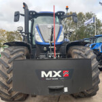 Front view of a blue New Holland tractor with oversized front tires and a black protective plow, displaying the MX Multimass 1200 logo on the ballast block in a dealership lot.