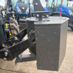 Close-up of a heavy black metal implement hitch with bolts and pin, attached to farming equipment, blue tractors blurred in the background.