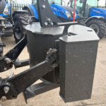 Close-up of a heavy black tow hitch and hydraulic linkage on a farm implement, with blue New Holland tractors in the background at a yard.