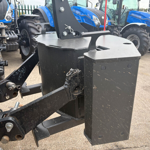 Close-up of a heavy black tow hitch and hydraulic linkage on a farm implement, with blue New Holland tractors in the background at a yard.