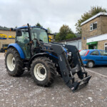Blue New Holland tractor with front loader in a parking lot outside a farm equipment shop; sign and brick building in the background.