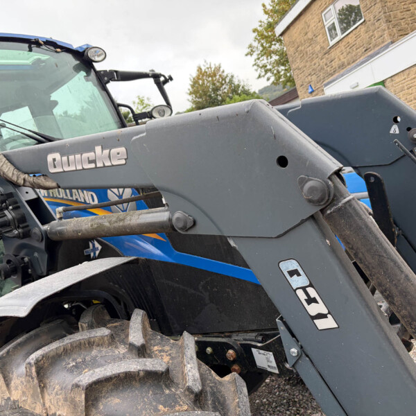 Close-up of a muddy blue tractor with a large gray loader attachment, parked beside a brick building.