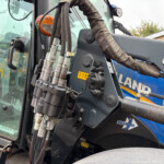 Close-up of hydraulic hoses and levers on a blue construction machine, with a worn protective sleeve and warning labels nearby.
