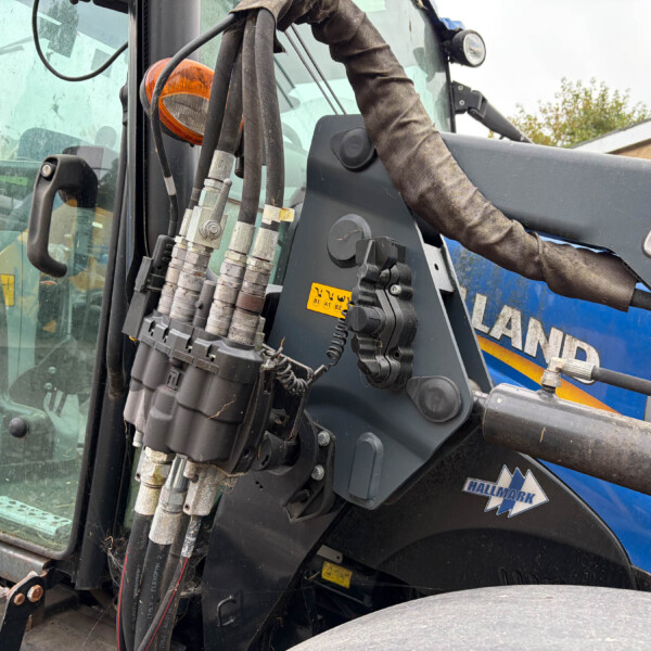 Close-up of hydraulic hoses and levers on a blue construction machine, with a worn protective sleeve and warning labels nearby.