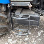 Close-up of a rusted tractor hitch with a grated footstep, hoses nearby and a muddy tire to the left.