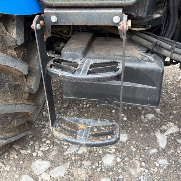 Close-up of a rusted tractor hitch with a grated footstep, hoses nearby and a muddy tire to the left.