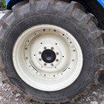 Close-up of a large muddy tractor tire with a white rim, dirt ground, and parked vans in the background.