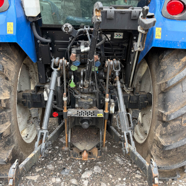 Rear view of a blue tractor showing the hydraulic hitch assembly with hoses, levers, and linkages, muddy tires on either side.