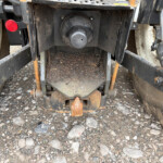 Rear view of a dirty trailer hitch with a circular, capped coupling in the center, rusty metal frame, red reflector on the left, and gravel ground.