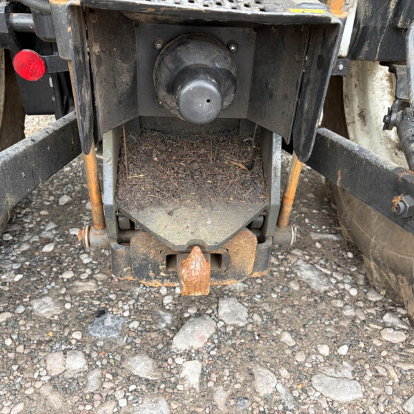 Rear view of a dirty trailer hitch with a circular, capped coupling in the center, rusty metal frame, red reflector on the left, and gravel ground.