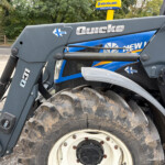 Close-up of a gray Quicke loader arm with a large muddy tire and a blue New Holland tractor in the background