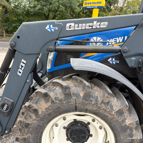 Close-up of a gray Quicke loader arm with a large muddy tire and a blue New Holland tractor in the background