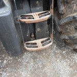 Close-up of a rusted metal step between tractor wheels on a dirt surface, showing heavy wear and grime.