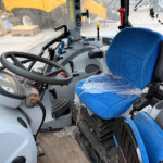 Cab interior of a construction vehicle with blue upholstered seat, steering wheel, and multiple levers; dusty windshield shows outdoor equipment and pallets outside