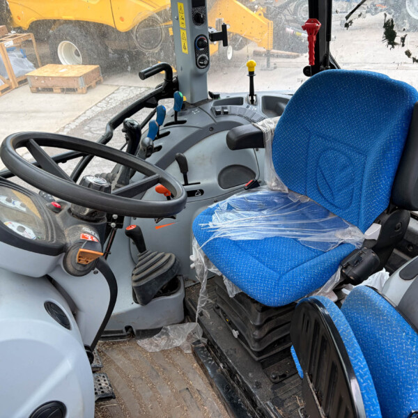 Cab interior of a construction vehicle with blue upholstered seat, steering wheel, and multiple levers; dusty windshield shows outdoor equipment and pallets outside