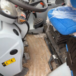 Interior of a dusty vehicle cab showing the steering wheel, pedals, and a blue fabric seat covered in plastic wrap.