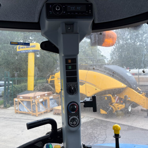 Inside the cab of a construction vehicle, showing the central dashboard with buttons, levers, and a rugged windshield with dirt and a yellow machine outside