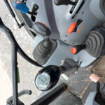 Close-up of a heavy equipment control panel with blue-grip levers, black levers, and an orange dial on a gray metal surface.
