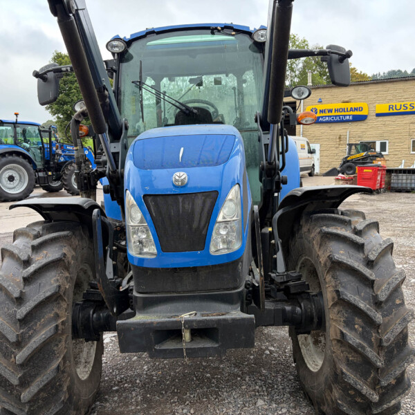 Blue tractor front view in a lot with other tractors, New Holland Agriculture sign on a building in the background