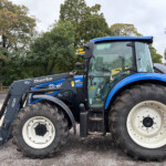 Blue agricultural tractor with a front loader attachment parked in a gravel lot, large tires and trees in the background.